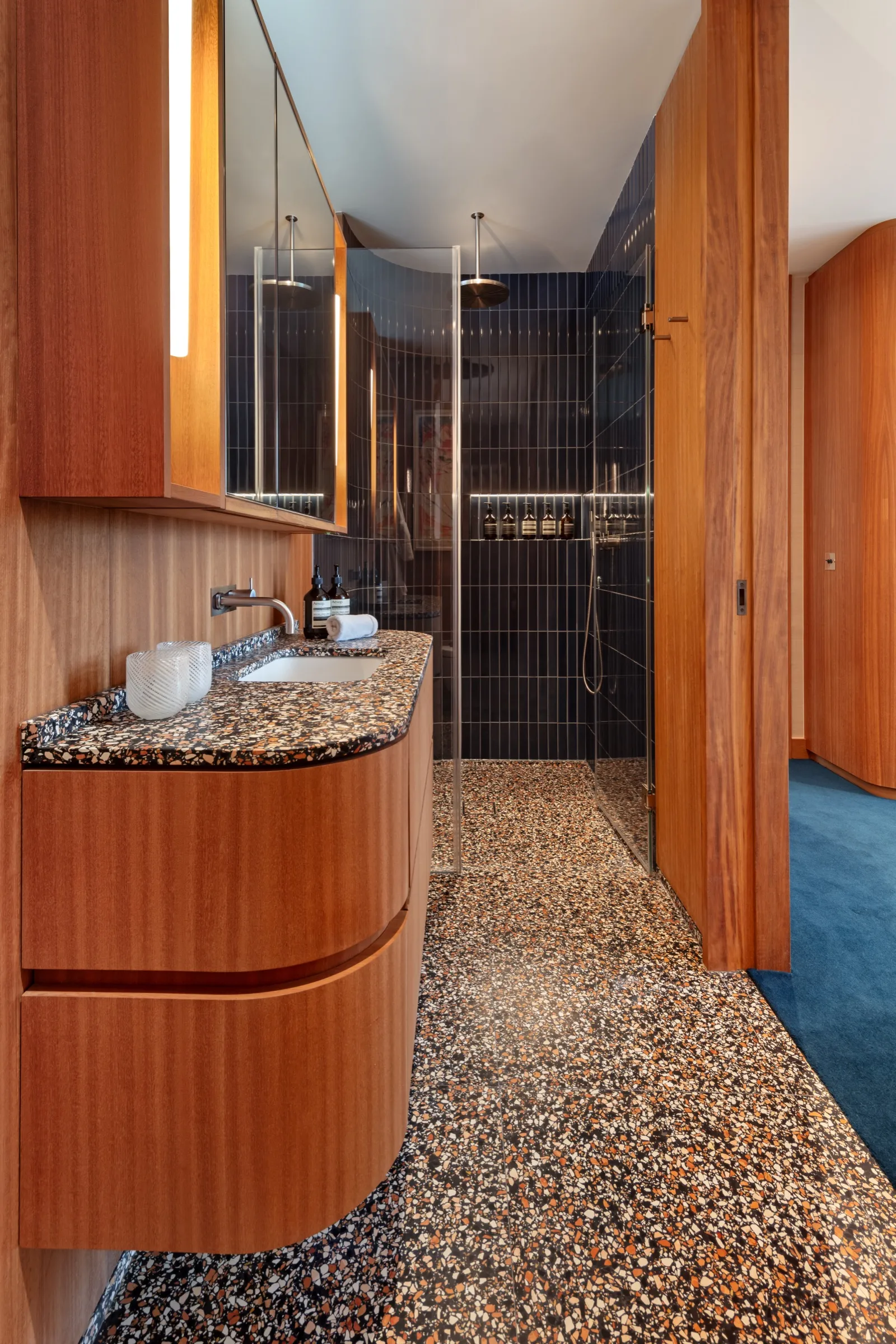 Curved timber vanity with terrazzo floor and dark tiled shower